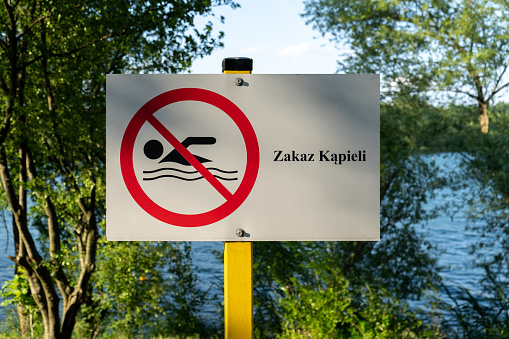 No Swimming sign post plaque, with water in background. No swim area signage pillar, mounted on the shore of lake in Poland. Text in Polish Zakaz kąpieli, means Swimming prohibited.