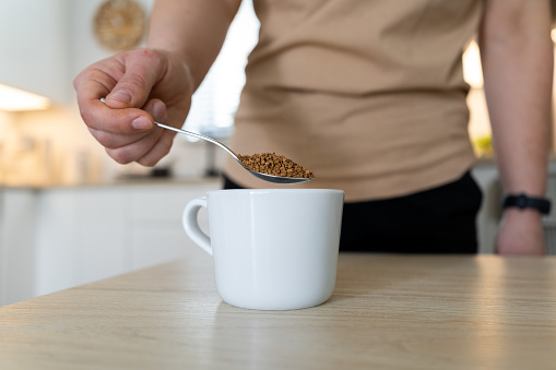 Woman pouring instant coffee granules into a white cup with a spoon. Girl with one spoonful of granulated coffee beans in a kitchen.
