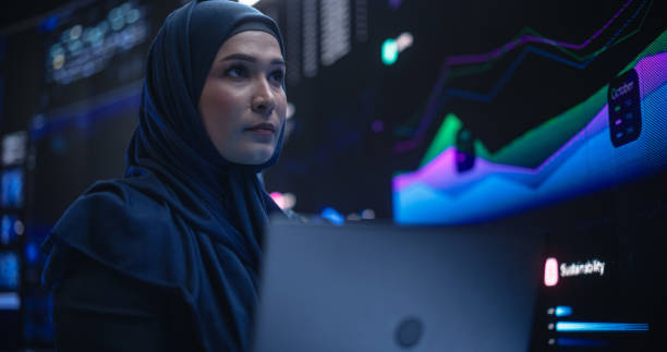 istock Muslim Female Data Center IT Engineer Standing in a Room with Statistics and Charts Settings on a Digital Screen. Cloud Computing Expert Uses Laptop for Servicing the System in Cyber Security Facility 1553010002