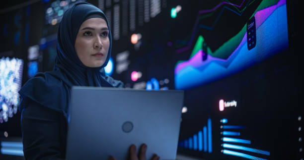 istock Muslim Female Data Center IT Engineer Standing in a Room with Statistics and Charts Settings on a Digital Screen. Cloud Computing Expert Uses Laptop for Servicing the System in Cyber Security Facility 1553010022