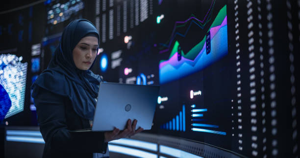 istock Muslim Female Data Center IT Engineer Standing in a Room with an AI Neural Network Settings on a Digital Screen. Cloud Computing Expert Uses Laptop for Servicing the System in Cyber Security Facility 1553010544