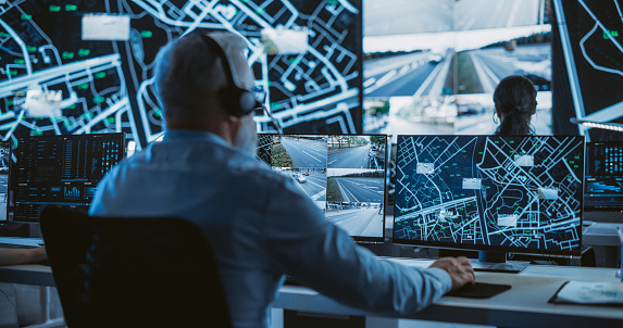 Senior Specialist in a Dark High-Tech Monitoring Room, Concentrated on a Call and Computer Work. Fleet Manager Using Headphones with Built-In Mic for Communication with Delivery Vehicles