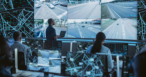 Male Software Engineers Working on Laptop Next to a Big Screen in a Modern Monitoring Office with Live CCTV Footage with Traffic Situation. Monitoring Room With Big Data Engineers Work on Computers
