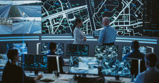 istock Two Multiethnic Software Engineers Talking Next to a Big Screen in a Modern Monitoring Office with Live CCTV Footage with Traffic Situation. Monitoring Room Big Data Engineers Work on Computers 1553020721