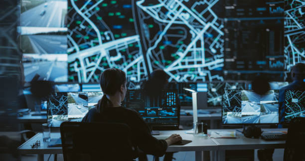 istock Establishing Shot: Dark Room with Multiethnic Specialists Working on Desktop Computers. Big LED Screen Shows Surveillance Satellite Data, City Map, and CCTV Traffic Footage Reports 1553021779