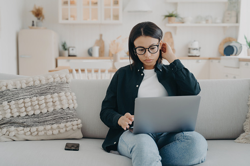Focused woman freelancer wearing glasses working on laptop sitting on couch at home. Concentrated young female check email, social networks, shopping online, browsing internet, using computer.