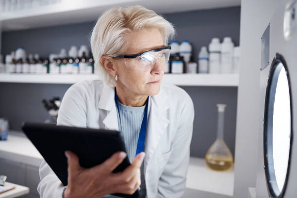 istock Scientist, tablet and senior woman with incubator for test, analysis or research study in laboratory. Technology, medical professional and serious science doctor check machine for biology experiment 1553096076