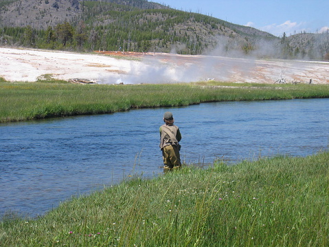 A young boy fishes at Yellowstone while a geyser lets of steam in the background. Coy space below and to both sides.