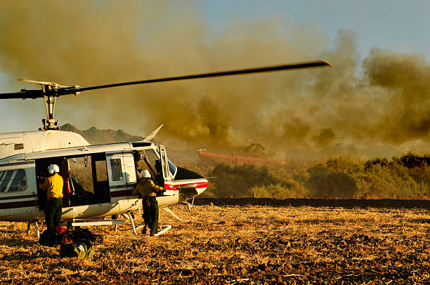 Two firefighters at helicopter in front of forest fire Helicopter team unloading the water bucket equipment as the pilot is briefed on details of the assignment. A small air tanker plane dropping retardant and smoke provide the backdrop to the scene. military tanker airplane photos stock pictures, royalty-free photos & images