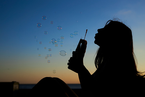 young girl with long hair blowing a bubble wand in clear blue sky sunset