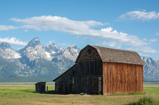 Rustic log barn has wooden roof and is in disrepair.  Field behind barn rolls across the distance to the mountains of Paradise Valley, Montana.