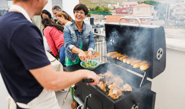 istock Happy friends cooking corncobs and chicken at barbecue rooftop dinner - Multigenerational people having fun during summer time eating and laughing together - Main focus on woman face 1553185727