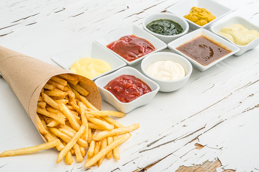 French fries wrapped in paper among different sauces in white saucers on the white wooden background with cracked paint. Top view.