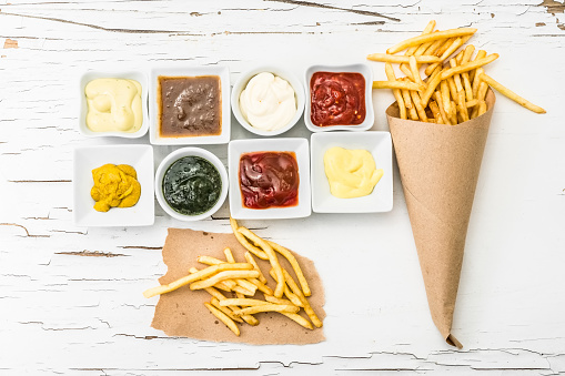 French fries on a piece of crafted paper with different sauces in white sausers on the white wooden background with cracking paint