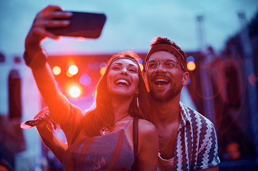 Cheerful couple taking selfie in front of a stage during summer music concert.