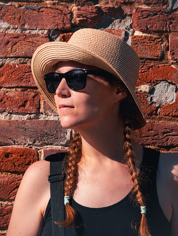 Young woman in straw hat portrait on red brick wall urban background. Millennial girl walking on summer city street with bright sunshine. Stylish confident authentic lady in black sunglasses outdoors.