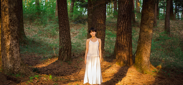 A short-haired Asian woman in a white dress standing in the twilight forest.