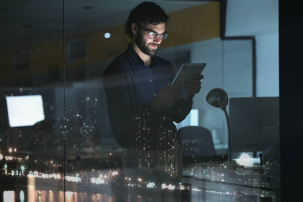 istock Business man, tablet and research in office, online browsing or internet scrolling by window with city lights at night. Technology, happiness and professional person with touchscreen for deadline. 1553289756