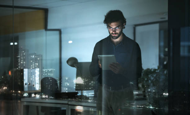 istock Business man, tablet and serious in office, online browsing or web scroll by window with city lights at night. Technology, working late and professional person with touchscreen for research deadline. 1553289771