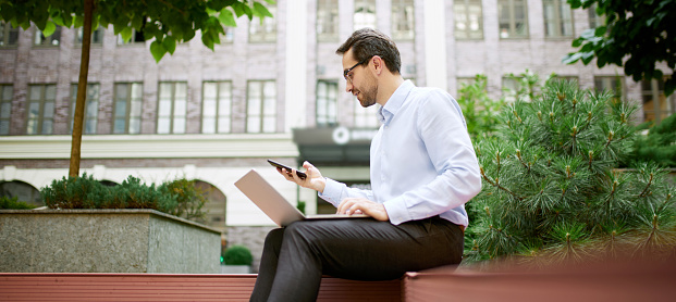 Young businessman sitting on bench outside of his office and working online with laptop and mobile phone. Concept of business, career development, ambitions, success, office lifestyle