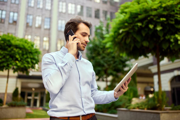 istock Young businessman standing outside office outdoors, looking on tablet and talking on mobile phone. Conversation 1553323261