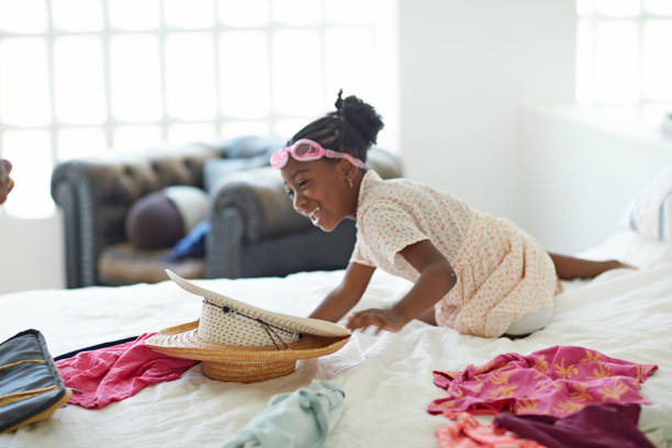 Laughing 6 year old Black girl sitting on bed in family home with swim goggles on her forehead and helping off-camera mother prepare for travel.