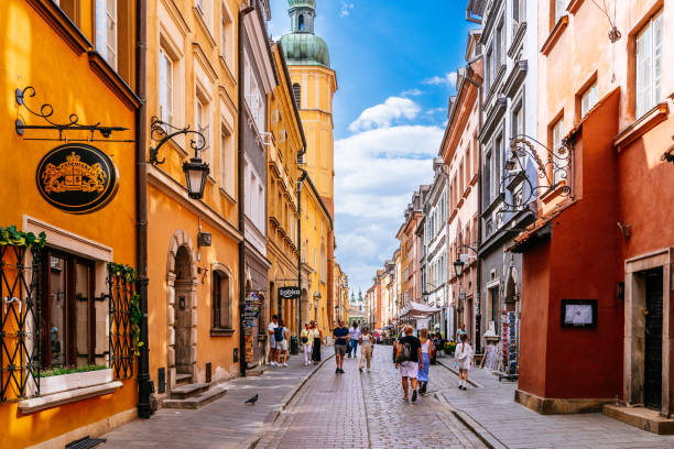A view of Piwna Street, a narrow street located within Warsaw's Old Town City Centre. The image captures the everyday scene of people walking along the street, engaging in leisurely activities. The architectural features of the surrounding buildings contribute to the distinct character of the area. The sky above is characterized by a blue hue and scattered clouds, creating a picturesque backdrop for the urban landscape. The photo provides a glimpse into the vibrant atmosphere of Piwna Street, offering a glimpse of daily life in this bustling section of Warsaw's historic city center.