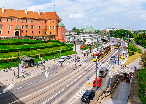 A view of Aleja Solidarności Street from Castle Square (Plac Zamkowy). The street is bustling with activity as people wait for public transport. Cars can be seen moving along the street. In the frame, a tram approaches a tram station, highlighting the public transportation system in the area. The photo offers a glimpse of the lively urban environment, showcasing the daily movements of people and vehicles on Aleja Solidarności Street.