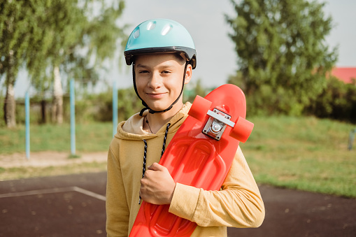 Portrait of happy schoolboy in blue helmet, holding red urban cruiser, skateboard in his hands on a sunny summer day in a skatepark. Beginner skateboarder