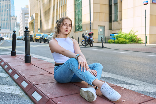 Young beautiful fashionable girl 19, 20 years old sitting on sidewalk, modern urban style. Natural blonde woman with blue eyes, curly hair looking at camera, European city, road background