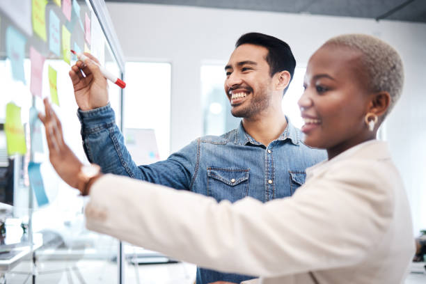 istock Happy creative people, writing and glass board for schedule planning, strategy or brainstorming at office. Man and woman in teamwork for startup project plan, sticky note or tasks at the workplace 1553413337