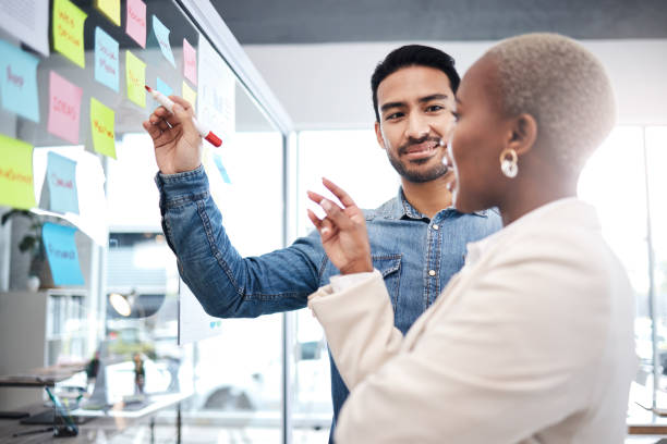 istock Creative people, writing and meeting on glass board for schedule planning, strategy or brainstorming at office. Man and woman in teamwork for startup project plan, sticky note or tasks at workplace 1553413384