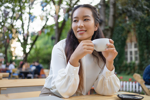 Young Beautiful Smiling Asian Woman in White Blazer With Cup Of Coffee Sitting in Coffee Shop
