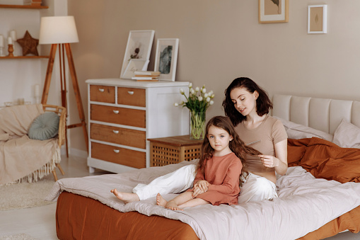Beautiful young mother combing her little daughter hair while sitting in bed in the morning. Bedroom interior with furniture made of natural materials. Concept of motherhood, household chores, family.