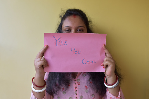 An Indian woman holding paper with text yes you can while standing in front of the wall. Copy space. Selective focus.