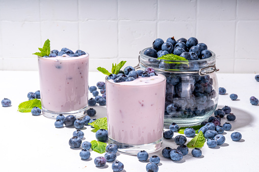 Blueberry yogurt, smoothie or milkshake on portioned glasses with fresh blueberries decor on it, and a lot of fresh berries on white kitchen table background copy space