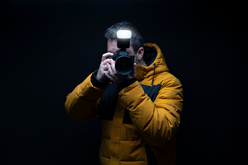 Portrait of a photographer in a yellow winter coat taking a photo with flash with a black background in studio