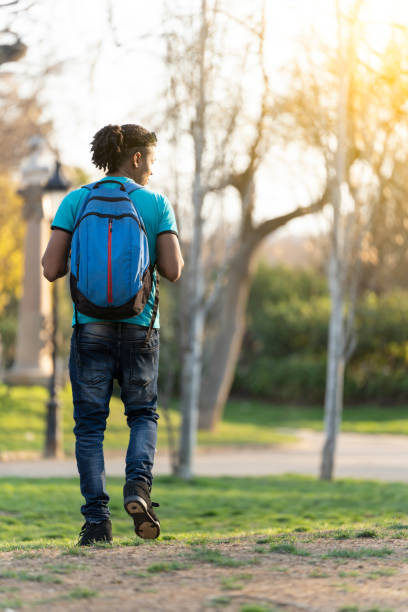 Young black student with curly hair is walking around the park with some trees on the background
