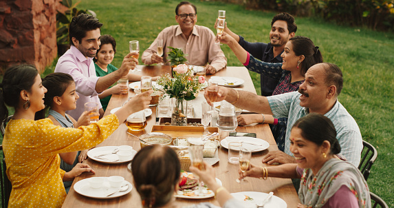 Happy Indian Family Celebrating: Group of People Gathered Together at the Table Raise Glasses To Make a Toast and Clink Glasses. Big Family Garden Party Celebration and Festivities