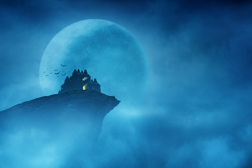 Light glows from a haunted house perched at the edge of a cliff in front of a large moon on a dark and spooky Halloween night.