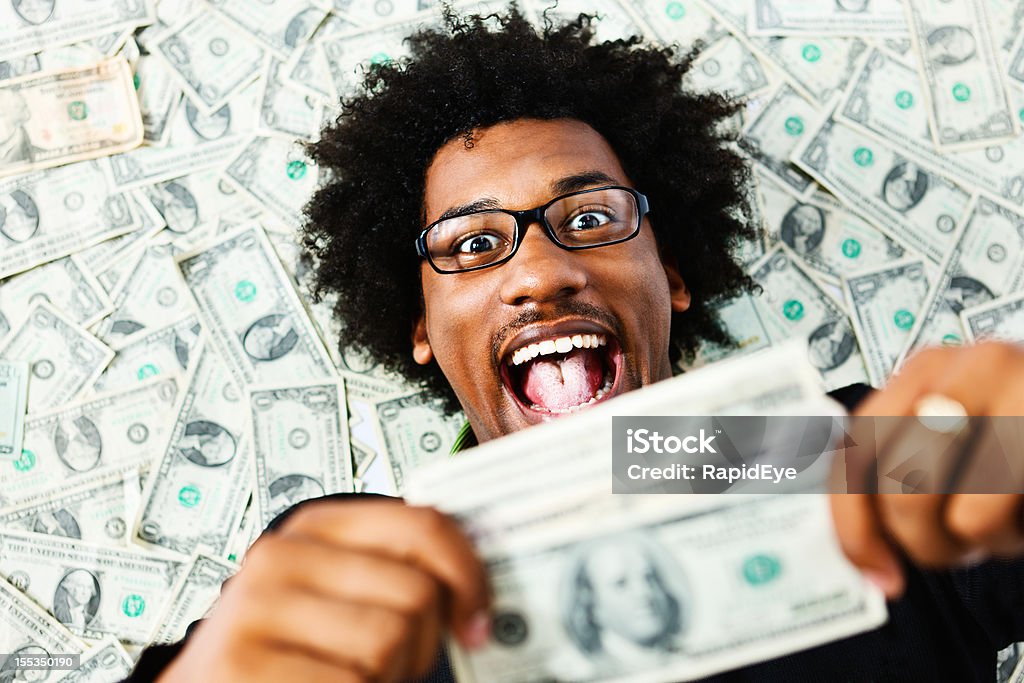 Overjoyed young man surrounded by US dollars A young man, with glasses and a wild afro, lies on a pile of US dollars, clutching a bundle of them,  and looking totally delighted at his good fortune. Currency Stock Photo