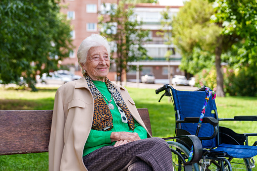 In a vibrant, leafy urban park, a cheerful elderly woman rests on a bench, basking in the sun after a wheelchair excursion.