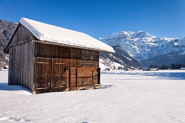 A high-angle view of a lonely wooden hut surrounded by a smooth, pristine expanse of deep white snow. This minimalist winter scene evokes a powerful sense of isolation, peace, and remote wilderness. The simple architecture of the timber shelter contrasts with the overwhelming scale of the snowy mountain environment