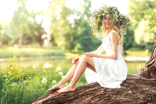 Portrait of a cute beautiful girl in a wreath and a white dress, sitting on a big tree in the forest. Ivan Kupala holiday.