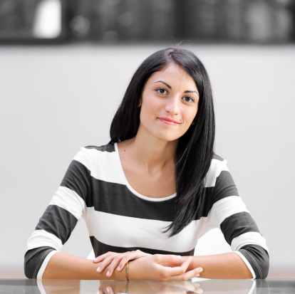 Square crop of a young woman sitting at a glass desk.