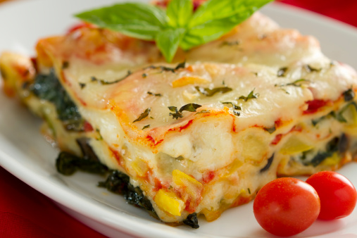 A serving of piping hot vegetable lasagna made with eggplant, tomato sauce, zucchini, summer squash, and Kalamata olives, combined with a creamy white sauce and topped with mozzarella cheese and chopped basal.