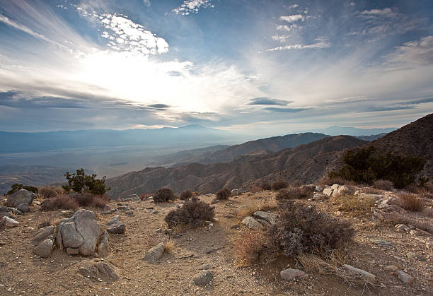 Grey and brown desert mountain range behind a red and gold plain of dried desert vegetation, with bright blue sky and fluffy clouds.