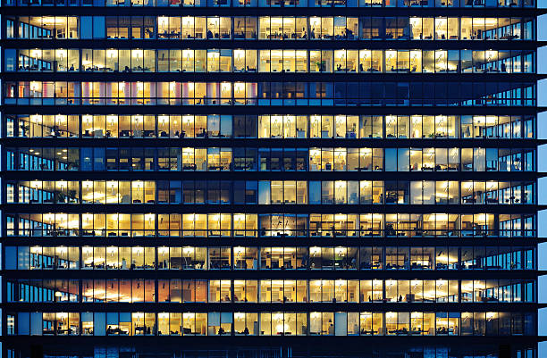 Workers working late. Office windows by night. Lots of people working late. Employees seen as silhouettes against their brightly lit offices with large windows. Building framed by the "blue hour" evening sky office building stock pictures, royalty-free photos & images