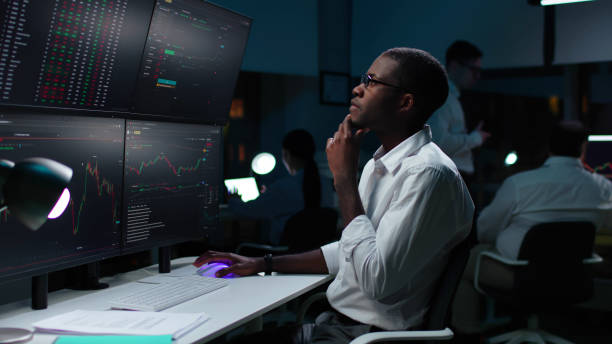 Financial analyst working on computer with multi-monitor workstation with real-time stocks Financial analyst working on a computer with multi-monitor workstation with real-time stocks. Pensive African-American businessman work in investment bank downtown office at night. stock brokers stock pictures, royalty-free photos & images