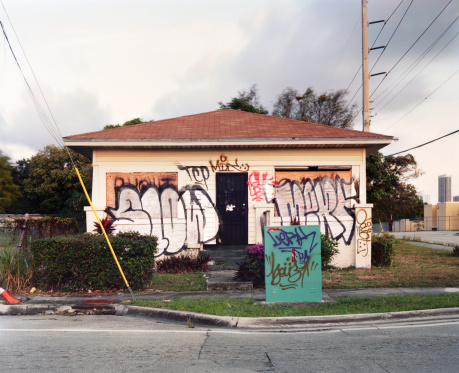 This is a horizontal, color photograph of a vacant Miami house. The windows are boarded up, covered with plywood and gang style tagging.